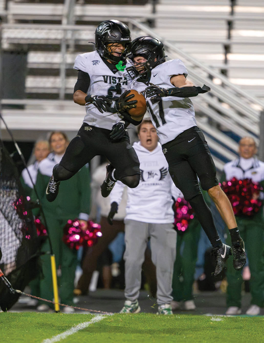 Two people in football uniforms jump in the air while holding a football on a field with cheerleaders and spectators in the background. Description generated by AI