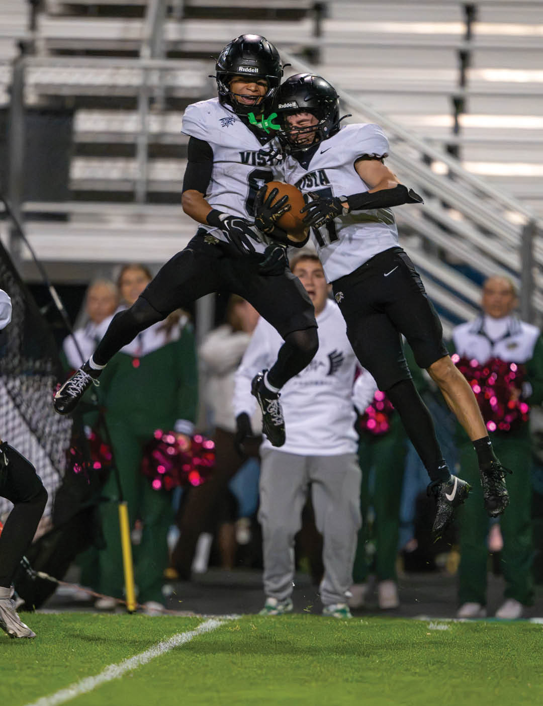 Two people in football uniforms jump in the air celebrating while one holds a football on a field with blurred spectators and cheerleaders in the background. Description generated by AI