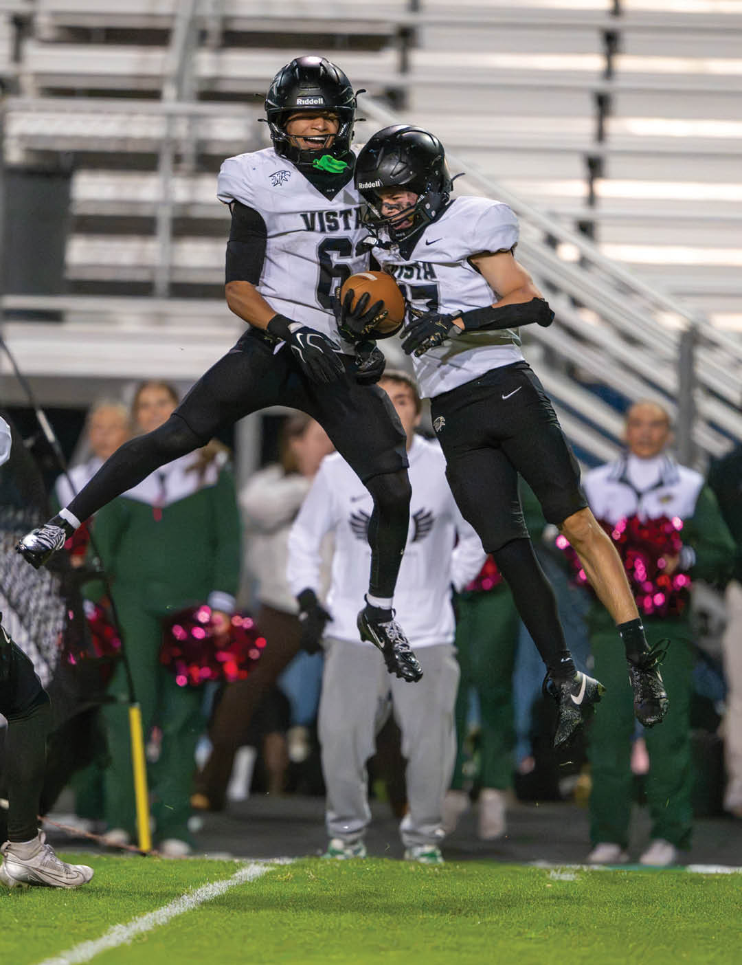 Two people in football uniforms jump in the air while one catches a football on a field with spectators in the background. Description generated by AI