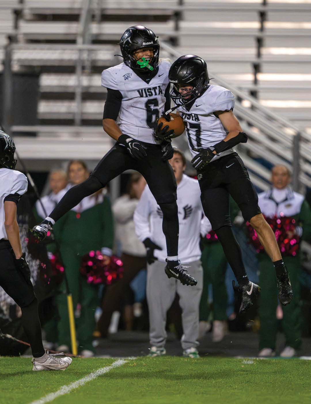 Two people in football uniforms jump in the air celebrating while holding a football on a field with spectators in the background. Description generated by AI