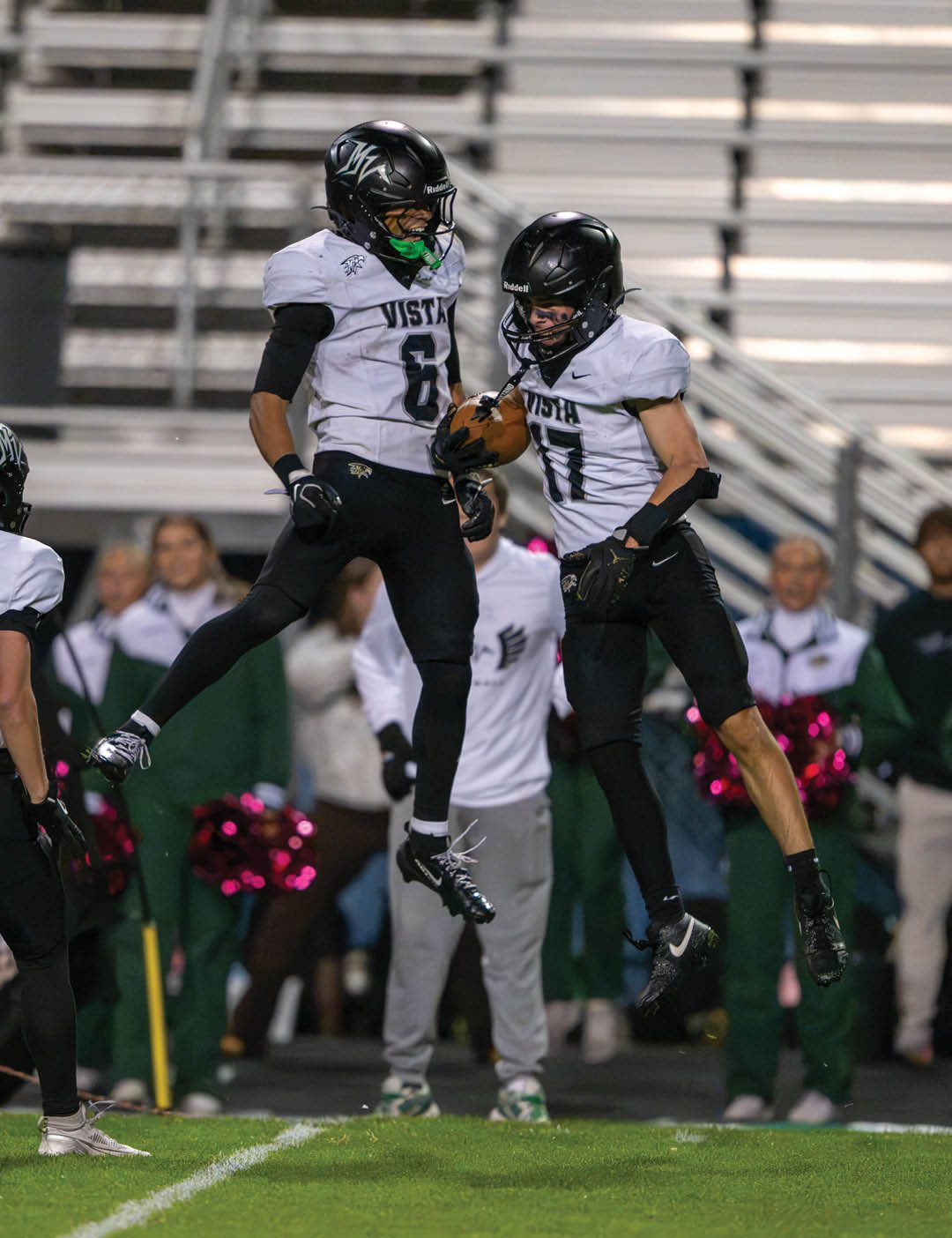 Two adults in football uniforms jump in the air celebrating on a field with cheerleaders and spectators in the background. Description generated by AI