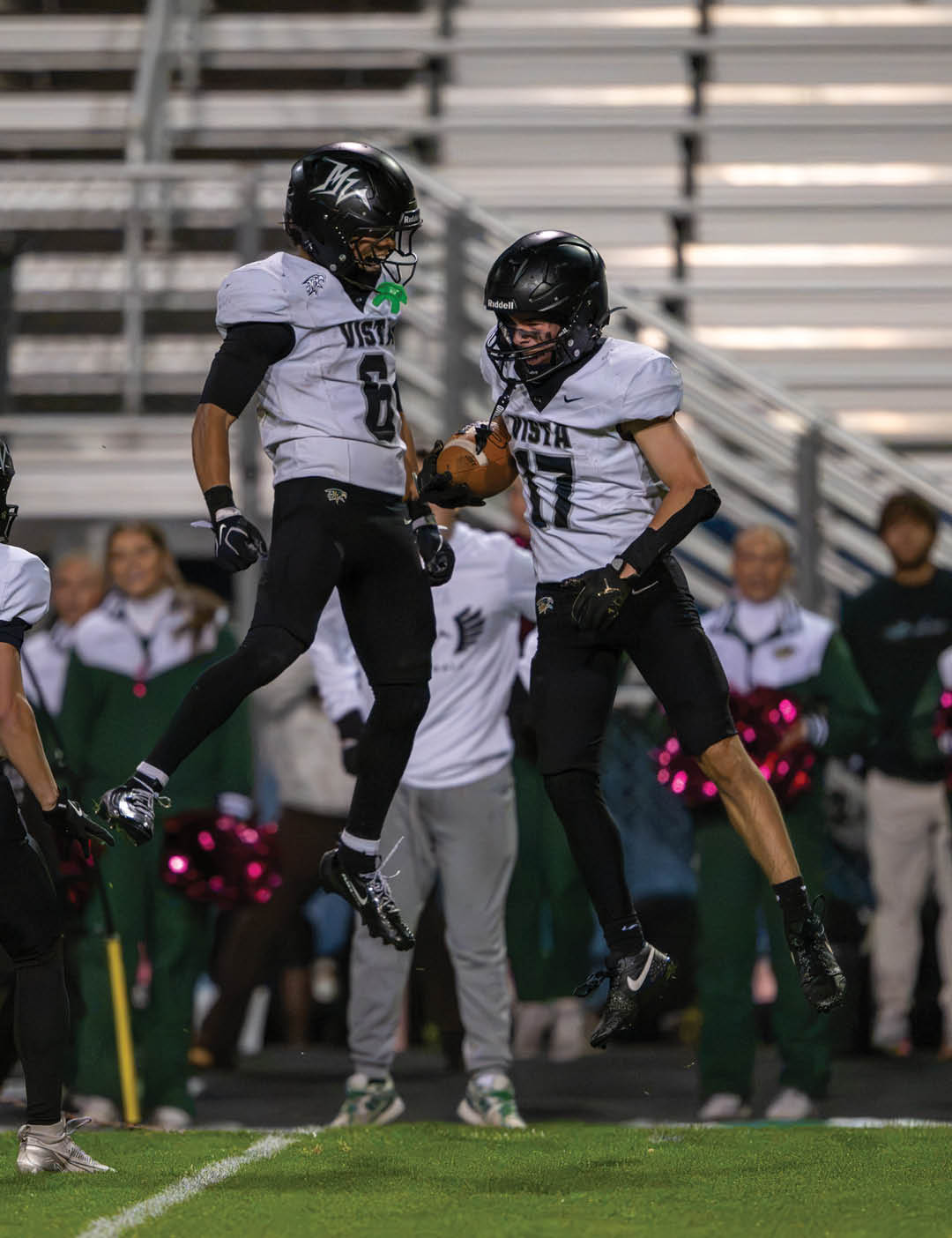 Two people in football uniforms jump in the air celebrating on a field with blurred spectators and cheerleaders in the background. Description generated by AI