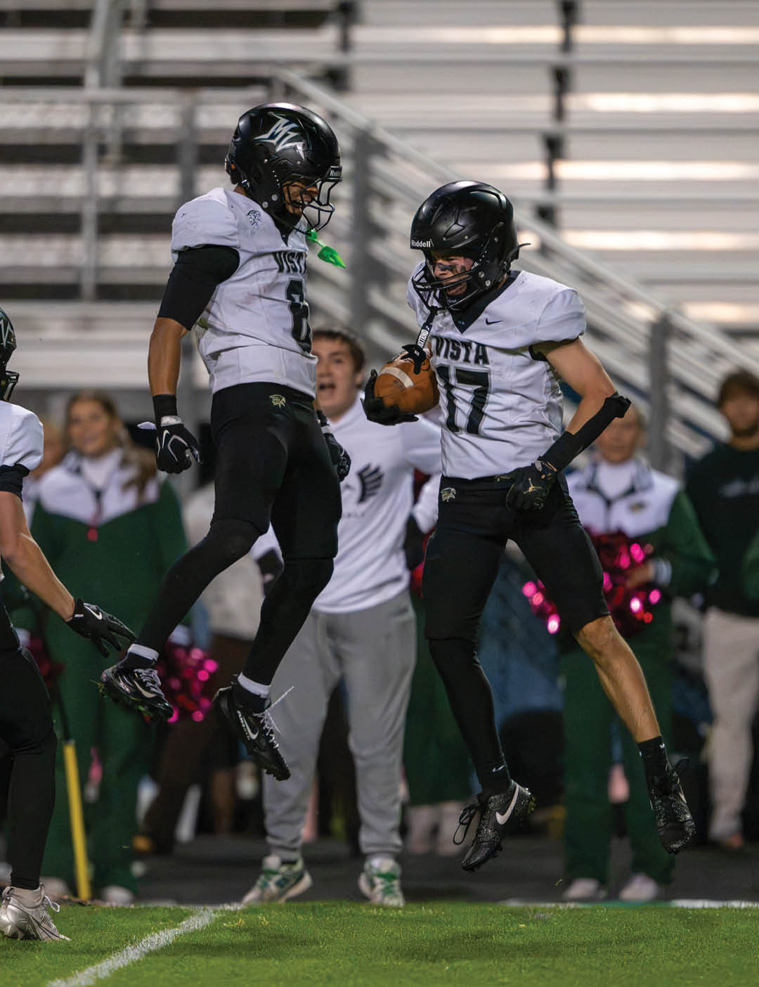 Two football players in black helmets and white jerseys jump in celebration on a field with spectators and cheerleaders in the background. Description generated by AI