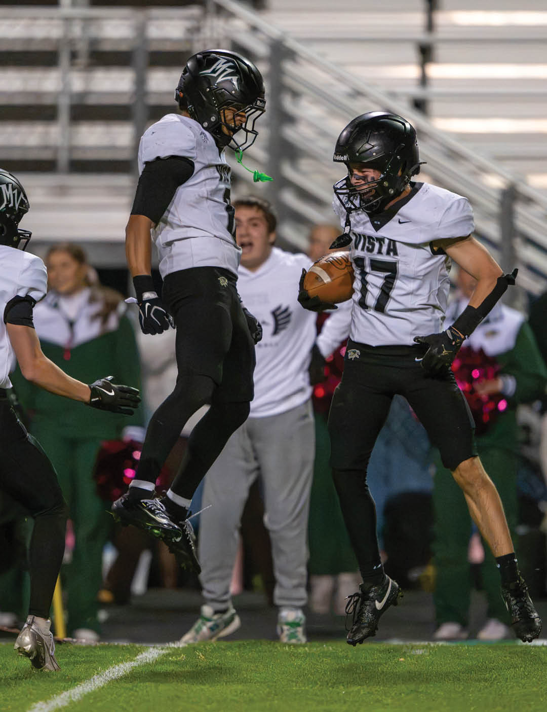 Two people in football uniforms celebrate on a field with one holding a football and the other jumping in the air while people watch in the background. Description generated by AI