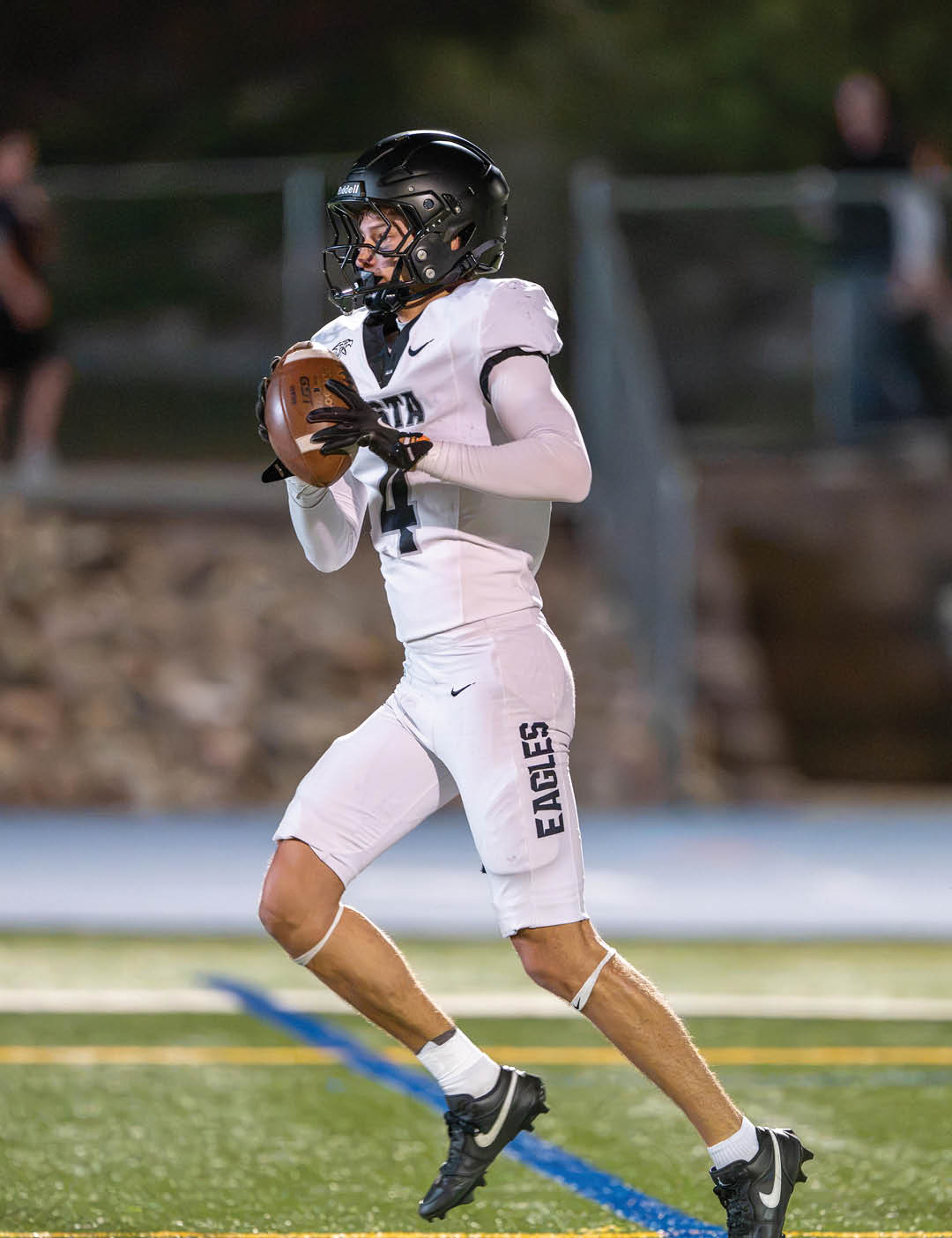 A person wearing a white football uniform with “EAGLES" on the pants and a black helmet is holding a football and appears to be preparing to throw it on a football field. Description generated by AI