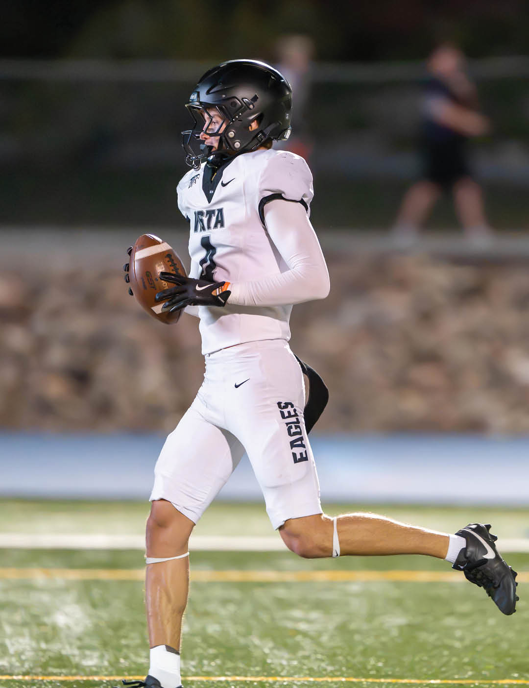 A person wearing a white football uniform with “Eagles" on the pants and a black helmet is running on a football field while holding a football. Description generated by AI