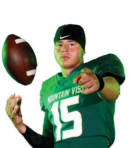 A person wearing a green football jersey with the number 15 and black athletic pants is tossing a football while pointing forward. Description generated by AI