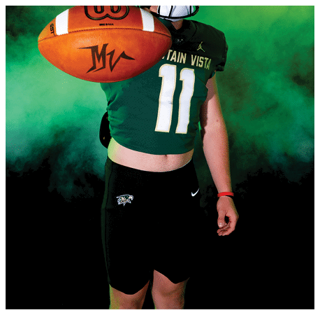 A person wearing a green football jersey with the number 11 and a black helmet is holding a football towards the camera against a smoky green and yellow background. Description generated by AI
