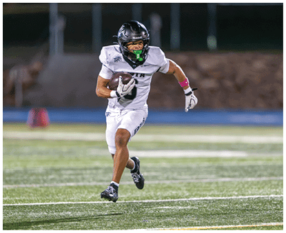 A person wearing a white football uniform and black helmet is running on a field while holding a football. Description generated by AI