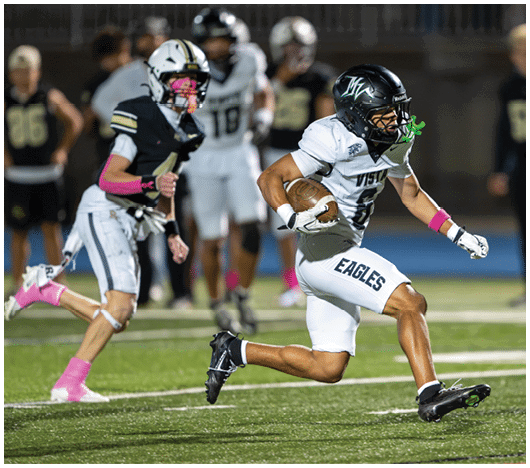 Two people in football uniforms are actively running on a field during a game, with one holding the ball and the other chasing behind. Description generated by AI
