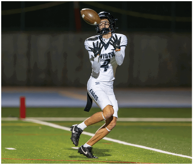 A person wearing a white football uniform and helmet is preparing to catch a football in mid-air on a green field. Description generated by AI