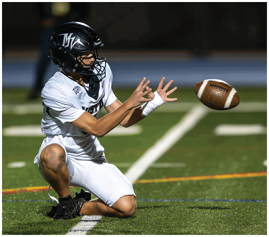 A person wearing a black helmet and white sports uniform is kneeling on a football field with arms extended, preparing to catch a football in midair. Description generated by AI