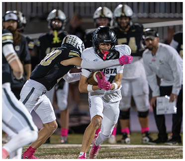 A group of people in football uniforms are actively playing on a field with one person in white running with the ball while others in black attempt to tackle. Description generated by AI