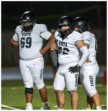 Three people in white football uniforms and black helmets stand on a field, appearing focused and ready for action. Description generated by AI