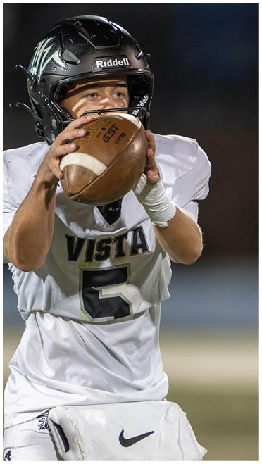 A person wearing a black helmet and white football uniform with the number five is preparing to throw a football on a field. Description generated by AI