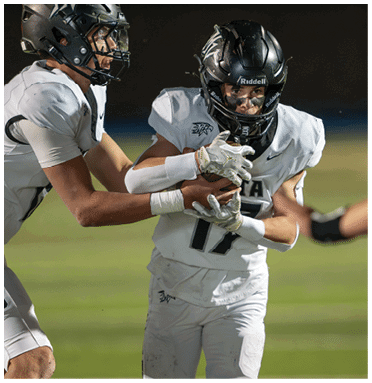 Two people in football uniforms are on a field with one handing off the ball to the other while a third person reaches out toward them. Description generated by AI