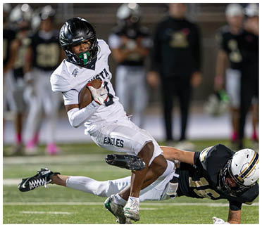 A person in a white football uniform with “EAGLES" on the pants is running with the ball while another person in a black football uniform is attempting to tackle them on a football field. Description generated by AI