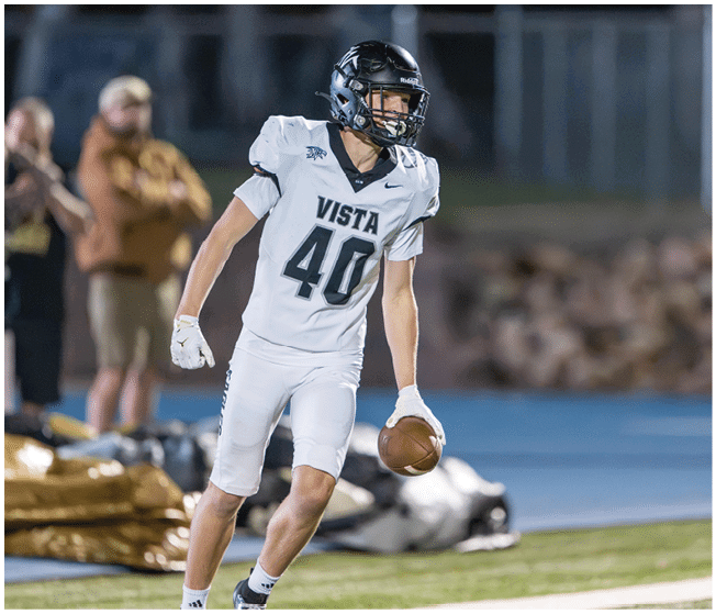 A person wearing a white football uniform with the number 40 and the word Vista is holding a football and standing on a field with blurred spectators in the background. Description generated by AI