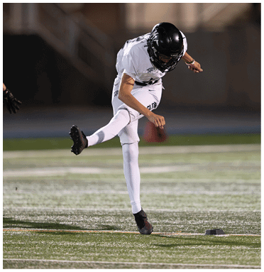 A person in a white sports uniform and black helmet is kicking a football on a field while another person in similar attire is partially visible nearby. Description generated by AI