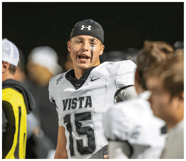 A person wearing a football uniform with the number 15 and the word Vista is talking to two other people in similar uniforms on a sports field at night. Description generated by AI