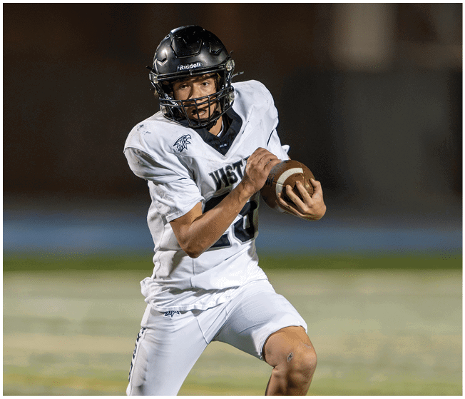 A person wearing a white football uniform and black helmet is holding a football and appears to be preparing to throw it on a field. Description generated by AI