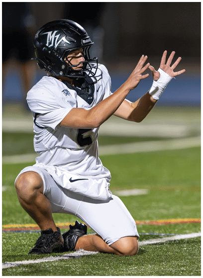 A person wearing a white football uniform and black helmet is kneeling on a field while reaching out to catch a football in midair. Description generated by AI