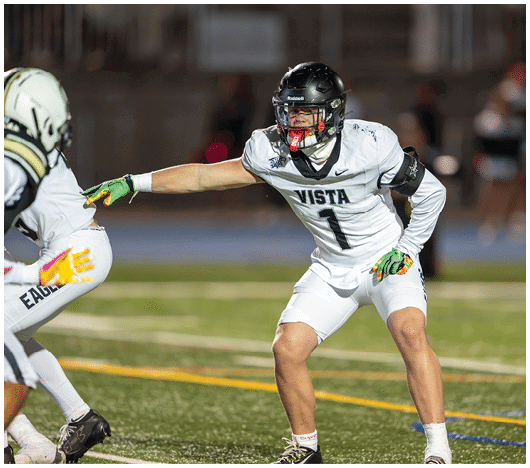 Two people in football uniforms are on a field with one person reaching out to the other while both appear focused and ready for action. Description generated by AI