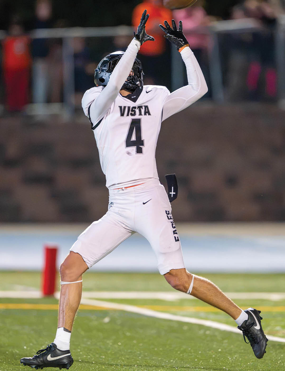 A person wearing a white football uniform with the word Vista and number 4 is reaching up to catch a football on a field. Description generated by AI