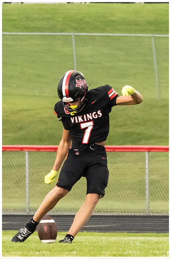 A man wearing a black and red jersey with the number 7 on it is kicking a football. Description generated by AI