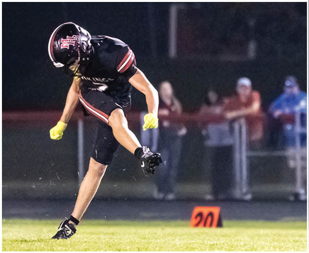 A football player in a black jersey with red trim is kicking a football. Description generated by AI