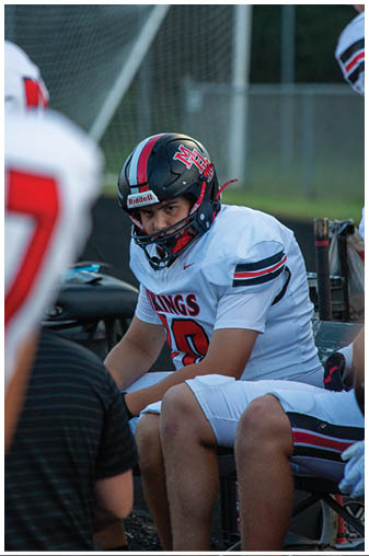 A football player wearing a helmet and number 18 sits on a bench. Description generated by AI