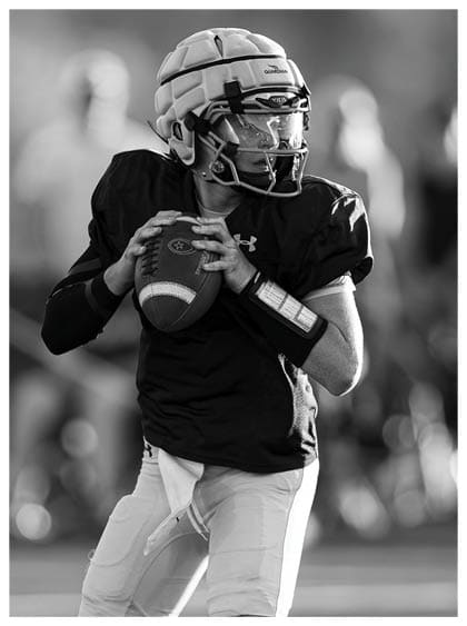 A person wearing a helmet and sports uniform is holding a football and preparing to throw it on a blurred outdoor field. Description generated by AI