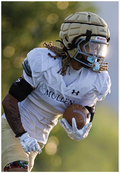 A person wearing sports gloves and athletic shorts is running while holding a football on a grassy field. Description generated by AI
