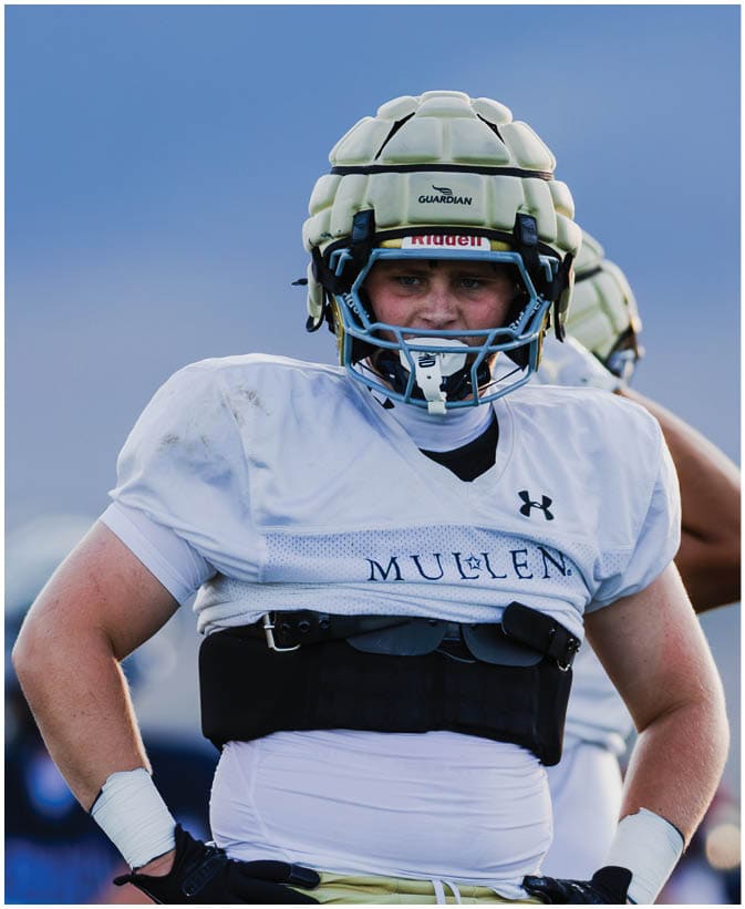 A person wearing a white football jersey with the word “MULLEN" and a padded helmet stands with hands on hips against a blurred outdoor background. Description generated by AI