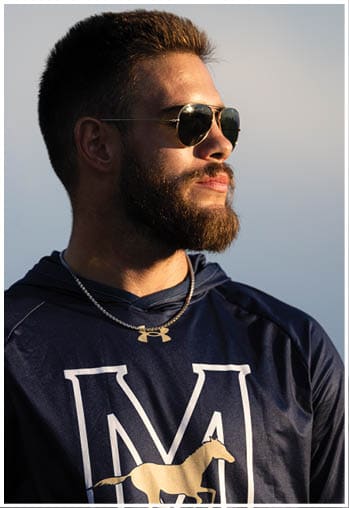A person wearing sunglasses and a dark blue hoodie with a large letter M and a horse graphic on it is looking to the side in natural light. Description generated by AI