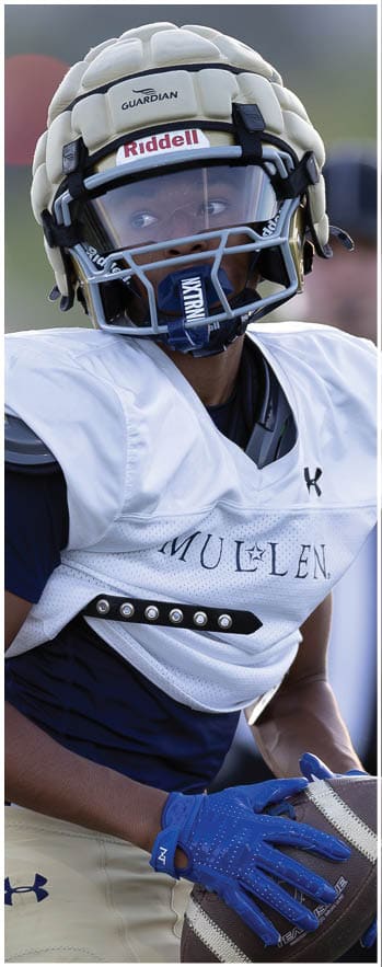 A person wearing a navy blue and white sports jersey with beige Under Armour football pants and blue gloves is holding a football. Description generated by AI