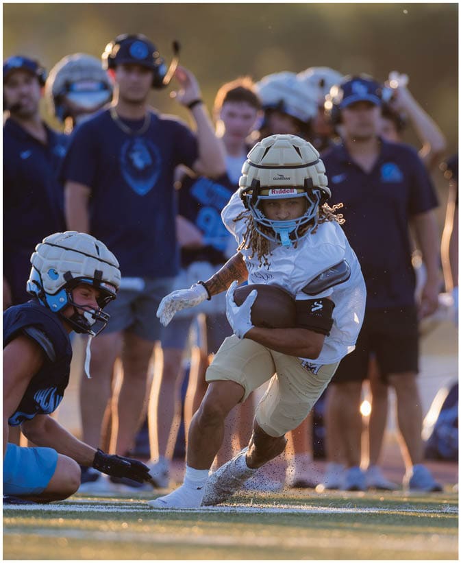 A person wearing a white football uniform and helmet is running with a football on a field while several people in blue uniforms and helmets stand in the background. Description generated by AI
