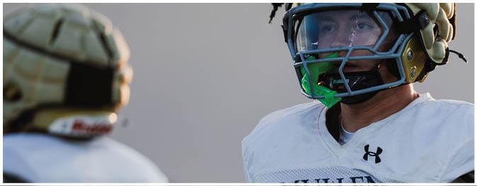 Two people wearing football helmets and white jerseys are facing each other, with one person in clear focus and the other blurred in the foreground. Description generated by AI