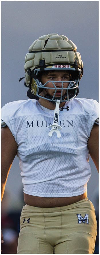 A person wearing a padded helmet and white sports jersey stands on a field with an arm raised in the background. Description generated by AI