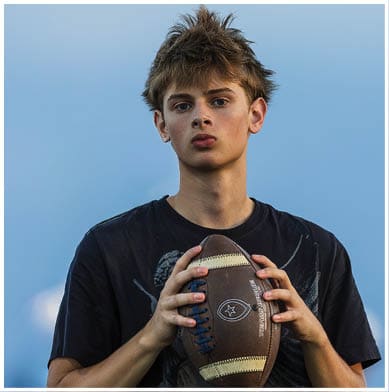 A person with tousled hair wearing a dark shirt is holding a football against a clear blue sky background. Description generated by AI