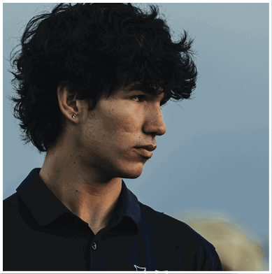 A person with curly hair wearing a dark collared shirt looks thoughtfully to the side against a blurred outdoor background. Description generated by AI