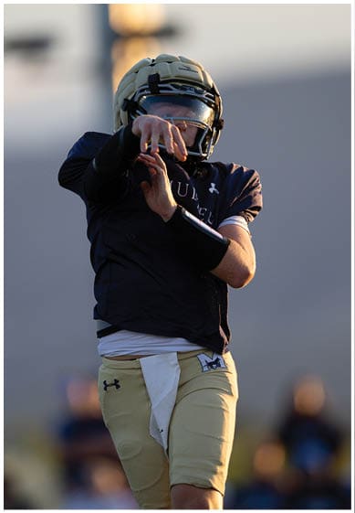A person wearing a helmet and sports uniform is preparing to throw a football on a field with blurred spectators in the background. Description generated by AI