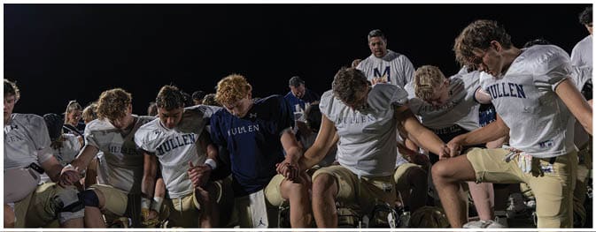A group of people wearing white sports jerseys are gathered closely together under a dark sky, with one person standing and appearing to speak to the others. Description generated by AI
