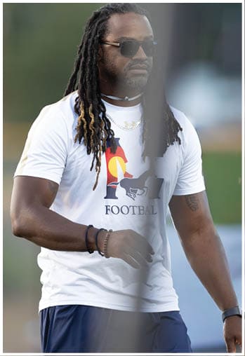 A person with long dreadlocks wearing sunglasses, a white graphic t-shirt, and a watch is walking outdoors near a blurred background. Description generated by AI
