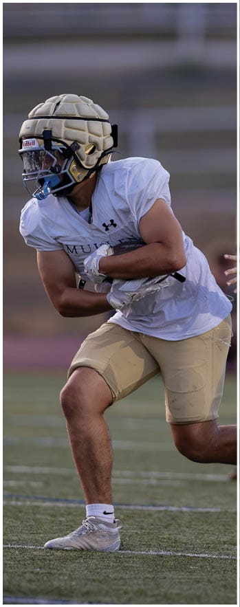 An adult wearing a white sports jersey, beige shorts, and a padded helmet is running on a football field while holding a football, with another person reaching out toward them. Description generated by AI