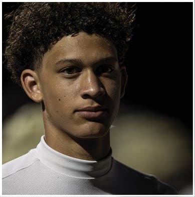 A person with curly hair wearing a white high-collar shirt is looking directly at the camera against a dark background. Description generated by AI