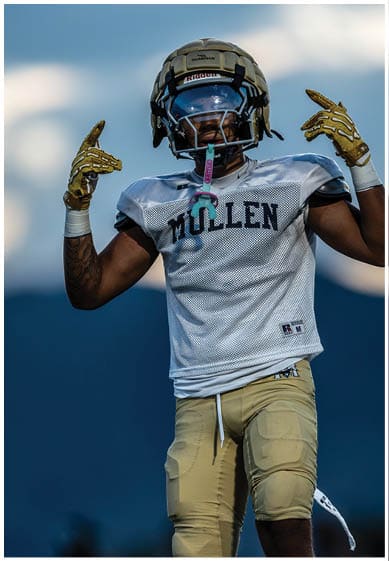 A person wearing a football helmet, visor, and gloves is raising one hand with a finger pointing upward against a blue sky background. Description generated by AI