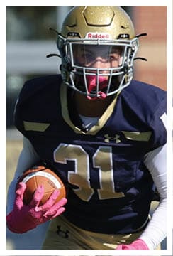 A person wearing a dark blue and gold football uniform with the number 21 and a helmet with a pink mouthguard is focused and ready for action. Description generated by AI