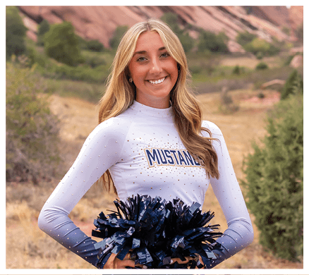 A person with long hair wearing a white and blue cheerleading uniform holding blue and black pom-poms stands outdoors with rocky hills and greenery in the background. Description generated by AI