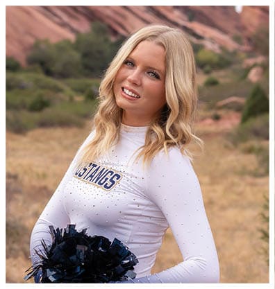 A person with long wavy blonde hair wearing a white cheerleading uniform with “MUSTANGS" on the front is smiling and holding dark blue pom-poms outdoors with a natural rocky background. Description generated by AI